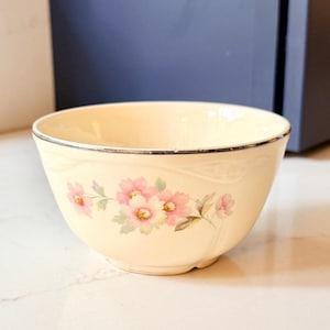 May include: Cream-colored ceramic bowl with a floral design. The bowl features a delicate pattern of pink flowers with yellow centers and green leaves. A thin silver-colored band trims the rim of the bowl. The bowl is likely vintage or antique.