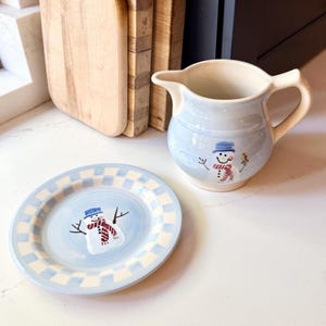 May include: A light blue ceramic pitcher and plate set with a snowman design. The pitcher has a handle and spout. The plate has a checkered border. The snowman has a blue hat, red scarf, and black twig arms. A charming winter-themed set.