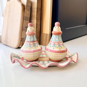 Pu&ograve; includere: Una coppia di saliera e pepiera in ceramica con vassoio abbinato dai bordi ondulati. I contenitori presentano un motivo dipinto a mano con forme geometriche e decorazioni floreali nei colori rosa, giallo, verde e blu su fondo crema.