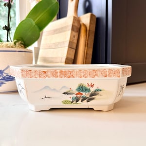 Może przedstawiać: Prostokątna biała ceramiczna donica z ręcznie malowaną sceną krajobrazu przedstawiającą góry, drzewa i mały czerwony budynek. Obrzeże ma pomarańczową i czerwoną ozdobną obwódkę. Donica ma około 25 cm długości.