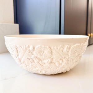 May include: Off-white ceramic bowl with embossed floral and leaf design. The bowl has a wide, rounded shape and a smooth, glossy finish. The embossed pattern covers the entire exterior of the bowl.