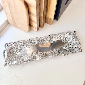 May include: A rectangular silver-toned metal serving tray featuring an intricate embossed border of grape clusters and vine leaves. The tray has a smooth, reflective center surface and integrated handles on both ends, showcasing a detailed botanical motif.