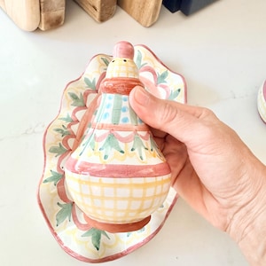Pu&ograve; includere: Un salino o pepaiolo in ceramica a forma di pera, caratterizzato da un motivo a griglia giallo dipinto a mano, fasce rosa e motivi a foglie verdi. Poggia su un vassoio ovale in ceramica abbinato con bordi smerlati e un disegno floreale.