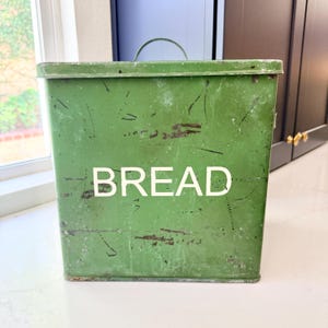May include: A square, green metal bread box with a distressed finish and visible surface scratches. The word BREAD is printed in large, white, sans-serif capital letters on the front. It features a flat lid with a small, arched metal handle.