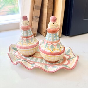 Pu&ograve; includere: Una coppia di saliera e pepiera in ceramica con un vassoio ovale abbinato dai bordi ondulati. Il set presenta un motivo dipinto a mano in rosa tenue, verde salvia e giallo, con linee a griglia, puntini e decorazioni a forma di foglia.