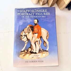 May include: A hardcover book titled Staffordshire Portrait Figures of the Victorian Era by P.D. Gordon Pugh. The blue cover features a photograph of a ceramic figurine depicting a man in a red shirt standing beside a white horse on a base.