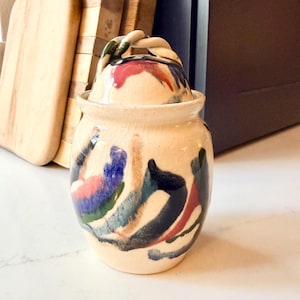 May include: A ceramic jar with a lid, featuring a cream-colored base and colorful abstract brushstrokes in blue, red, green, and black. The lid has a twisted handle. The jar is likely handmade and suitable for kitchen or home decor.