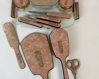 May include: A vintage vanity set with a pink and cream marbled design. The set includes a tray, jars, brushes, mirrors, and manicure tools. Each piece features a gold floral design. The set is arranged on a white surface.