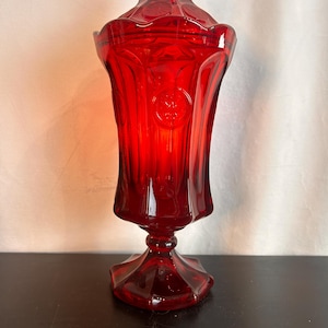 Puede incluir: Un bombonera de cristal rojo rubí con tapa. El plato tiene una base de pedestal y un cuerpo acampanado y acanalado. La tapa también es roja y tiene un adorno decorativo. El cristal tiene un acabado brillante.