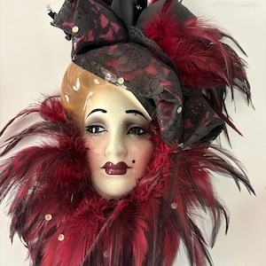 Puede incluir: Una máscara decorativa de cerámica con una cara pintada, maquillaje de ojos oscuro y labios color burdeos. La máscara está adornada con un sombrero de encaje y cinta negros, y un cuello de plumas rojas y negras. Pequeñas lentejuelas están dispersas.