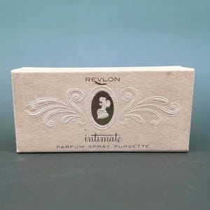 May include: A rectangular, off-white box with the Revlon logo and the word "Intimate" in cursive. The box features an oval design with a silhouette and decorative flourishes. The text "Parfum Spray Pursette" is printed below.