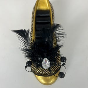 May include: A decorative gold-colored shoe with black accents. The shoe features a black feather and bead embellishment on the toe, with a large clear gem. The interior is gold with black satin ribbons.
