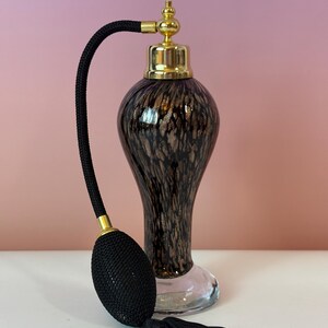 May include: An elegant vintage perfume bottle with a black bulb atomizer and tassel. The bottle has a gold-colored top and a dark brown and gold speckled design. The base is clear glass. The atomizer is black with gold accents.