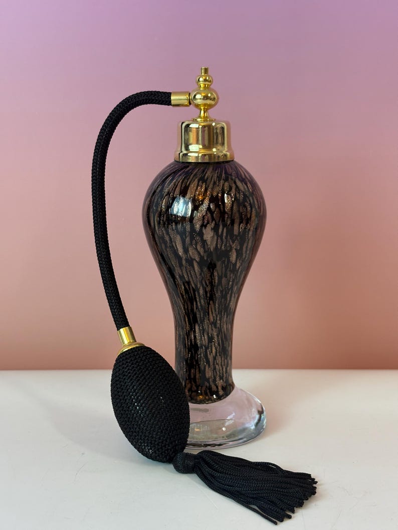 May include: An elegant vintage perfume bottle with a black bulb atomizer and tassel. The bottle has a gold-colored top and a dark brown and gold speckled design. The base is clear glass. The atomizer is black with gold accents.