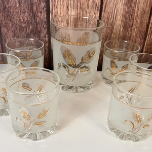 May include: A set of seven vintage frosted glass drinking glasses. The glasses feature a gold wheat stalk design and a decorative gold rim. The set includes a tall glass and six smaller glasses, all with a scalloped base.