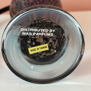 May include: A clear glass perfume bottle with a dark, speckled interior. The bottom of the bottle has a sticker that reads "DISTRIBUTED BY MAG PARFUMS" and "MADE IN TAIWAN". The bottle has a rounded shape.
