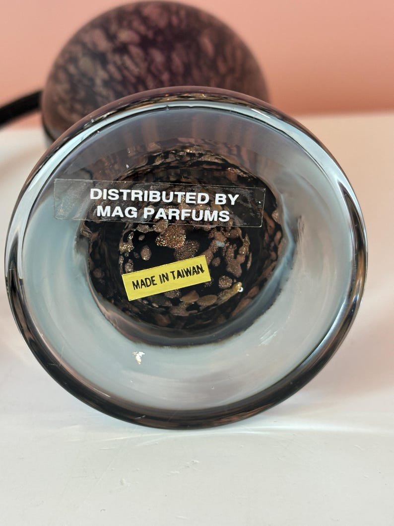 May include: A clear glass perfume bottle with a dark, speckled interior. The bottom of the bottle has a sticker that reads "DISTRIBUTED BY MAG PARFUMS" and "MADE IN TAIWAN". The bottle has a rounded shape.
