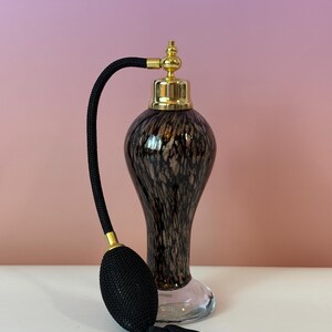May include: A vintage perfume bottle with a black bulb atomizer and tassel. The bottle is a speckled brown and gold color with a gold-colored cap. The base of the bottle is clear glass.
