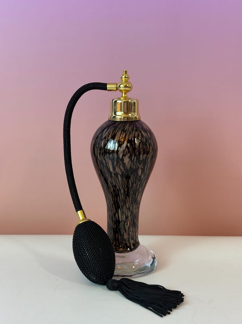 May include: A vintage perfume bottle with a black bulb atomizer and tassel. The bottle is a speckled brown and gold color with a gold-colored cap. The base of the bottle is clear glass.