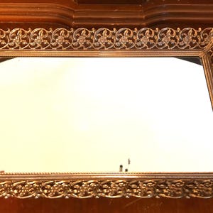 Op de afbeelding: Een rechthoekig dienblad met een reflecterende spiegelbasis en een versierde goudkleurige metalen rand. Het frame vertoont een ingewikkeld opengewerkt filigraanpatroon met herhalende bloemmotieven langs alle vier de randen van het dienblad.