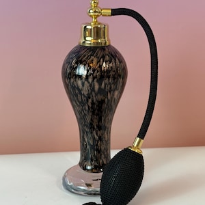 May include: An antique perfume bottle with a black and gold speckled design. The bottle has a gold-colored top and a black bulb atomizer with a matching tassel. The bottle sits on a clear, round base.