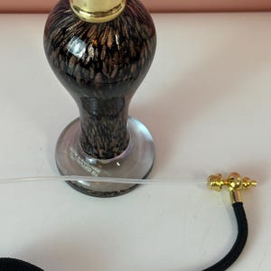 May include: A vintage-style perfume bottle with a black and gold patterned glass body, a gold-colored cap, and a black bulb atomizer with gold accents. The bottle has a clear glass base.