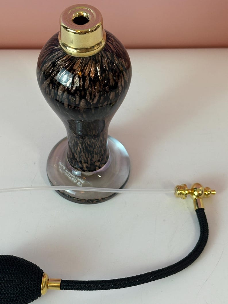 May include: A vintage-style perfume bottle with a black and gold patterned glass body, a gold-colored cap, and a black bulb atomizer with gold accents. The bottle has a clear glass base.