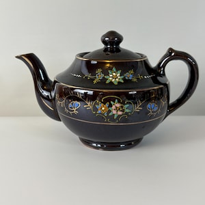 Könnte beinhalten: Eine dunkelbraune Keramik-Teekanne mit Deckel, Ausguss und Henkel. Die Teekanne ist mit goldenen Verzierungen und bunten Blumenmustern in Blau, Rosa und Grün verziert. Die Teekanne ist im Vintage-Stil.