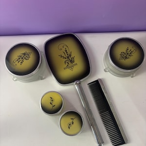 May include: A vintage vanity set comprising a hand mirror, comb, and jars. The set has a yellow and black floral design. The mirror and comb have silver-toned handles and frames. The jars have clear bases.