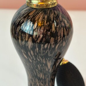 May include: A vintage perfume bottle with a black and gold speckled design. The bottle has a bulb atomizer and a gold-colored cap. The glass bottle has a rounded shape and a long neck. The atomizer is black.