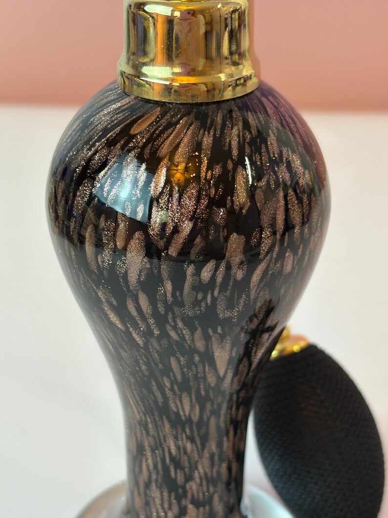 May include: A vintage perfume bottle with a black and gold speckled design. The bottle has a bulb atomizer and a gold-colored cap. The glass bottle has a rounded shape and a long neck. The atomizer is black.