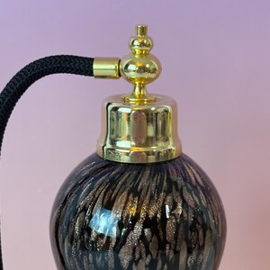 May include: A vintage perfume bottle with a dark purple base and gold flecks. The bottle has a gold-colored metal top and a black corded atomizer. The design is reminiscent of antique vanity items.
