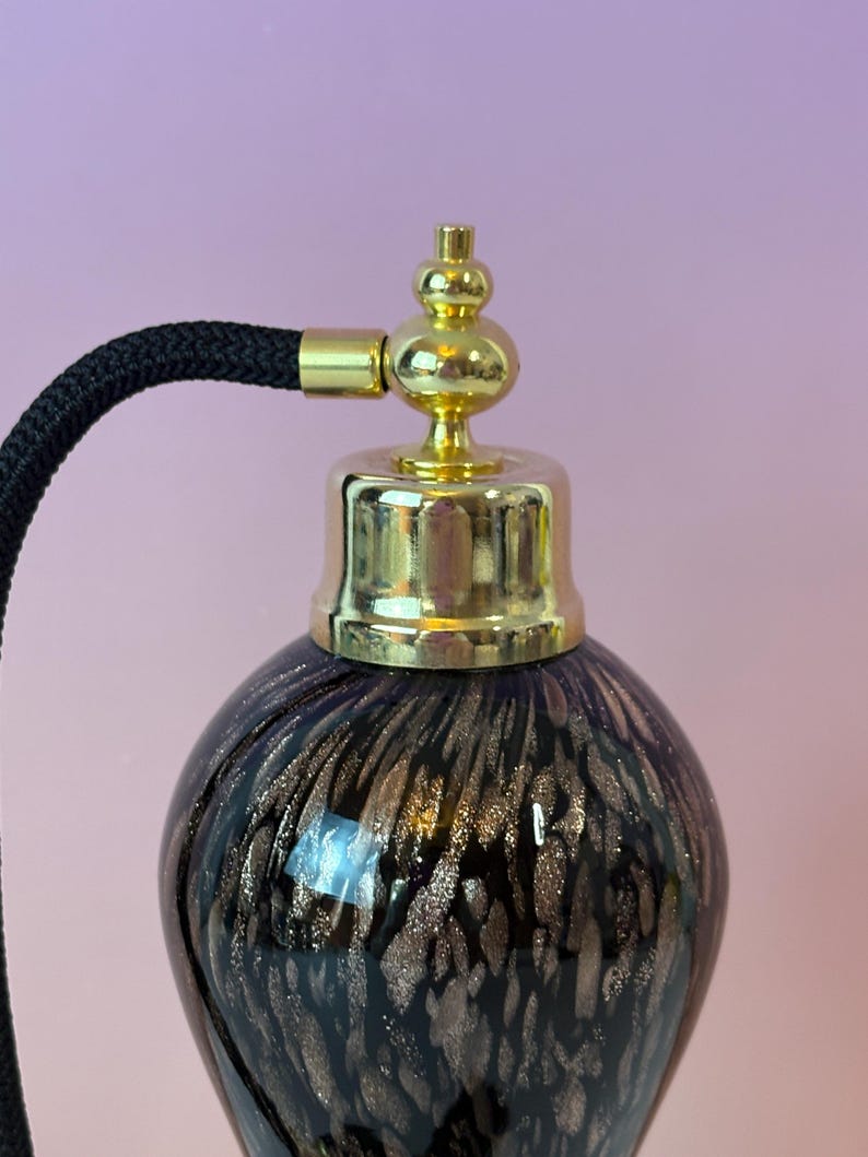 May include: A vintage perfume bottle with a dark purple base and gold flecks. The bottle has a gold-colored metal top and a black corded atomizer. The design is reminiscent of antique vanity items.