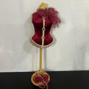 Op de afbeelding: Een rode fluwelen mannequin torso met gouden versieringen en groene edelsteen accenten. De mannequin staat op een houten standaard met een rode en gouden strik aan de basis.
