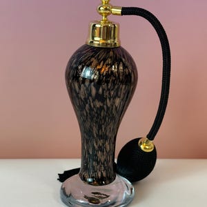 May include: An elegant perfume atomizer with a black and gold speckled glass body. It features a gold-colored top and a black bulb atomizer with a matching tassel. The base is clear glass.
