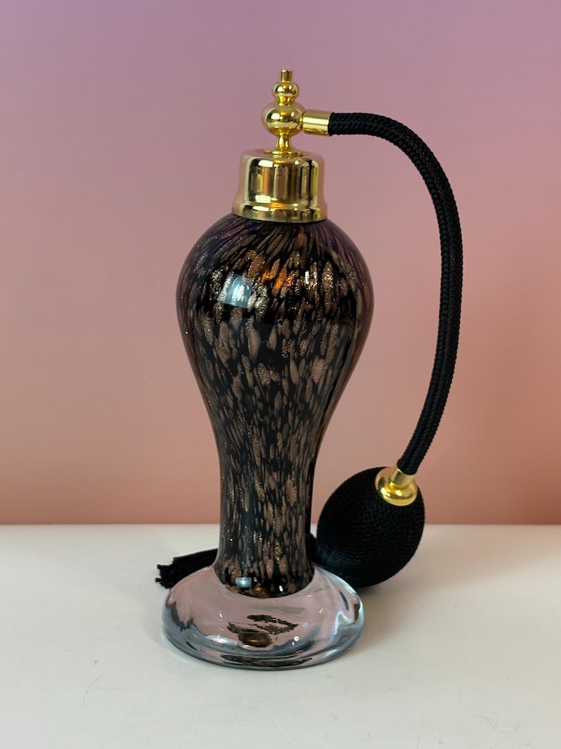 May include: An elegant perfume atomizer with a black and gold speckled glass body. It features a gold-colored top and a black bulb atomizer with a matching tassel. The base is clear glass.