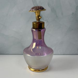 May include: A glass perfume bottle with a wide, frosted white base that transitions into a translucent purple neck. The bottle features a gold-toned metal collar and a decorative, textured, glittery purple cap shaped like a small flower.