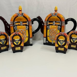 Może przedstawiać: Dwa ceramiczne czajniki w kształcie vintage'owych automatów muzycznych z pasującymi solniczkami i pieprzniczkami. Czajniki są brązowe, żółte i pomarańczowe z srebrnymi akcentami. Solniczka i pieprzniczka są brązowe i pomarańczowe.