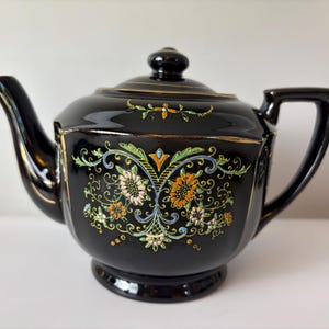 Könnte beinhalten: Schwarze Keramik-Teekanne mit Deckel und Henkel. Die Teekanne ist mit bunten Blumen- und Blattmotiven mit goldenen Verzierungen verziert. Ausguss und Henkel sind gebogen. Die Teekanne steht auf einer weißen Oberfläche.