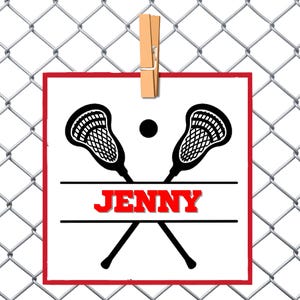 Pode incluir: Um cartão quadrado branco com uma borda vermelha, apresentando dois bastões de lacrosse pretos cruzados sob a palavra "JENNY" em vermelho. Um ponto preto está acima da palavra. O cartão é preso por um prendedor de roupa de madeira contra uma cerca de tela de arame.