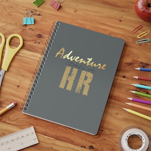 以下が含まれることがあります： 「Adventure Starts in HR」の文字が金色で書かれた、グレーのリング綴じノート。ノートは、ハサミ、鉛筆、クリップなどの学用品が置かれた木製の机の上にあります。ノートのサイズは約21.6cm×27.9cmです。