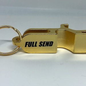 May include: A gold-coloured keychain bottle opener with the text "FULL SEND" in black. The opener has a ring for keys and a curved design for opening bottles. The background is a plain white surface.