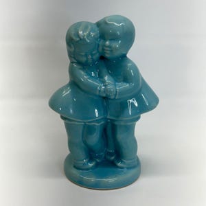 Peut inclure: Une figurine en céramique bleu clair brillant représentant deux enfants debout côte à côte, s'enlaçant. Les personnages sont sculptés avec des traits lisses et arrondis, vêtus de tenues simples avec des jupes évasées, le tout fini par un vernis monochrome réfléchissant.