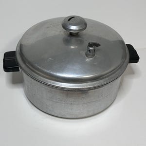 May include: A vintage aluminum pressure cooker with a domed lid, a pressure release valve, and black handles. The cooker has a circular shape and a shiny, metallic finish. The lid features a small, round knob.