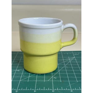 Könnte beinhalten: Eine Keramik-Tasse mit gestapeltem Design, die einen Farbverlauf aufweist. Der obere Rand ist weiß, geht in Gelb über und hat dann einen leuchtend gelben Sockel. Sie hat einen abgerundeten Griff.