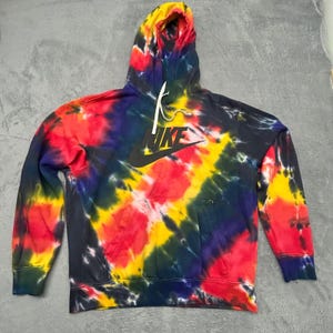 May include: A long-sleeve hooded sweatshirt featuring a vibrant tie-dye pattern in shades of red, yellow, blue, and black. The garment includes a front kangaroo pocket, a drawstring hood, and a large black Nike logo printed on the chest area.
