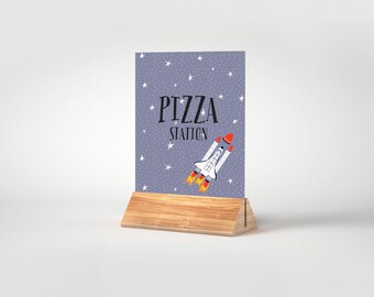 May include: A purple sign with white stars and a rocket ship graphic. The sign says "Pizza Station" in black text. The sign is on a wooden stand.