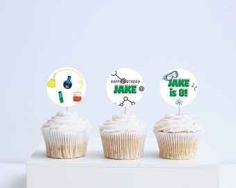 May include: Three cupcakes with white frosting topped with round science-themed cupcake toppers. The toppers feature green text that says "Happy Birthday Jake", "Jake is 8!" and a beaker, test tube, and flask.
