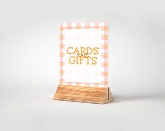 May include: A pink and white gingham sign with the text "CARDS and GIFTS" in orange. The sign is on a wooden stand.
