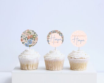 May include: Three cupcakes with pink and white frosting topped with round paper cupcake toppers. The toppers feature a floral pattern, a floral wreath with the text "Harper is one!" and a pink polka dot pattern with the text "Happy Birthday Harper".